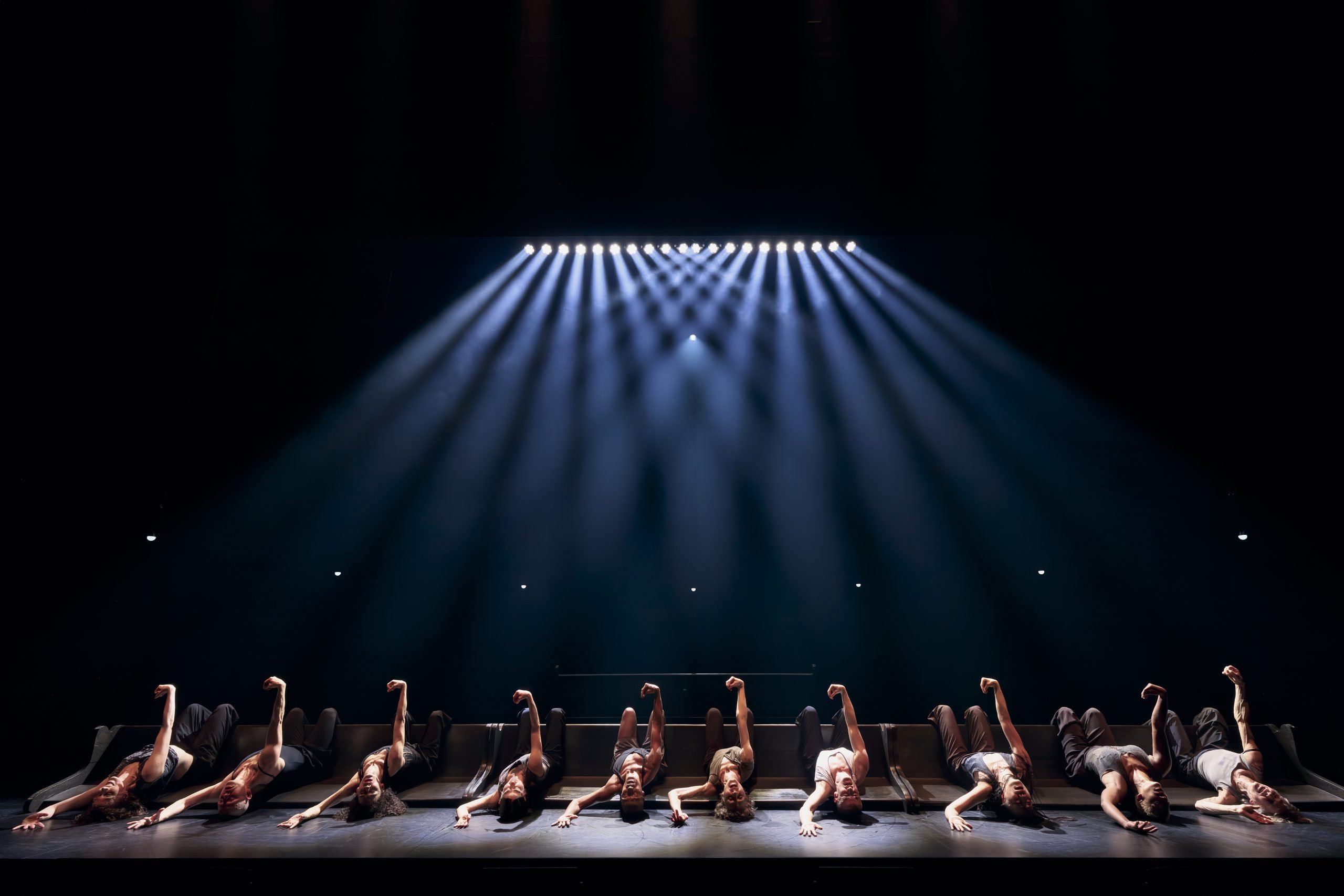 Dancers lying on their backs along the edge of a stage with arms raised, under blue spotlight beams.